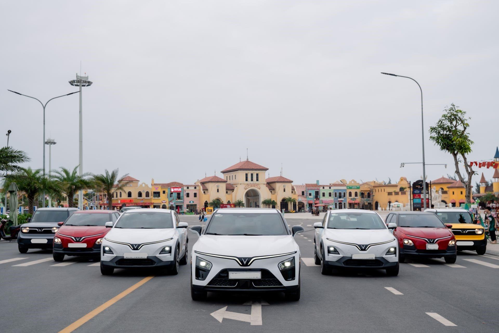 Đoàn xe VinFast gồm SUV điện trắng phía trước và các mẫu khác chạy trên đường, phía sau là khu nhà mái ngói.