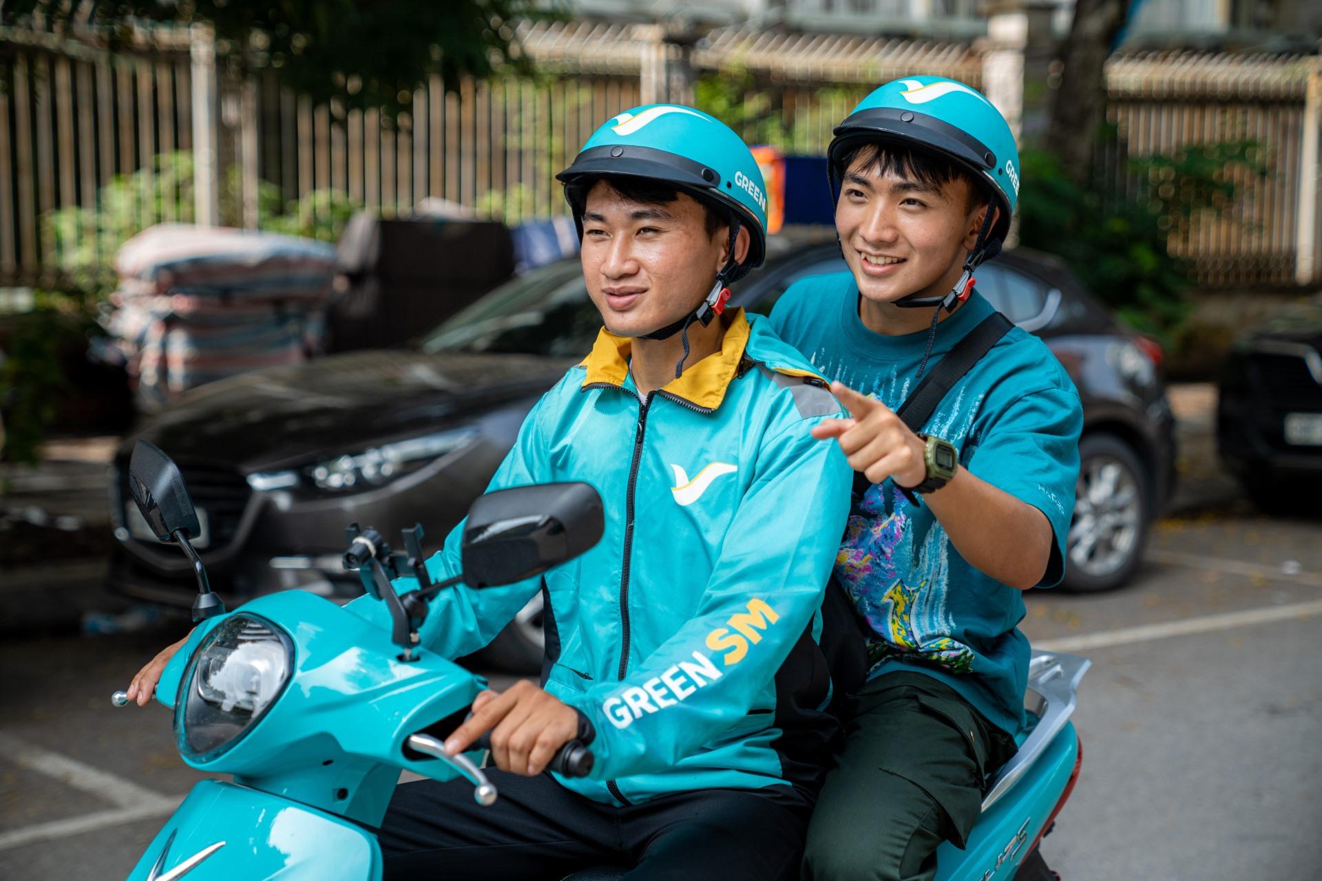 Hai người đội mũ bảo hiểm GREEN SM, mặc đồng phục xanh, chạy xe máy điện VinFast trên đường phố.